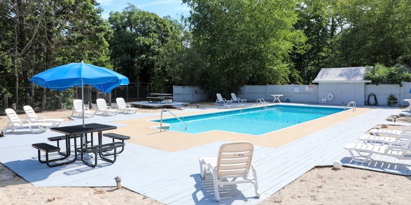 An inviting outdoor pool area with lounge chairs, a blue umbrella over a picnic table, and surrounded by trees, offering a relaxing space to enjoy the sun.