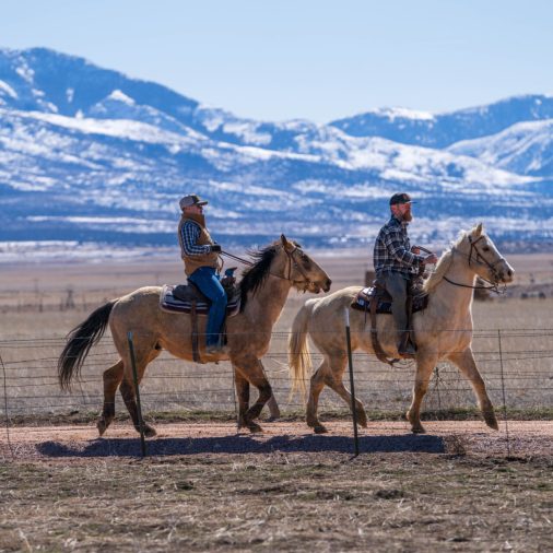 Two men riding horses in a wide, open field with snowy mountains behind.