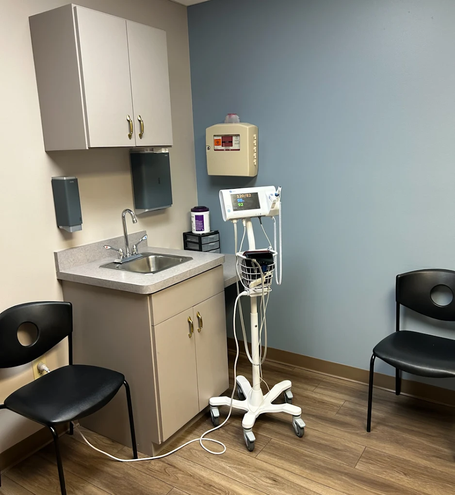 BrightView Health exam room with a vital signs monitor, sink and storage cabinets, medical supply dispensers, black patient chairs, blue accent wall, and wood-look flooring