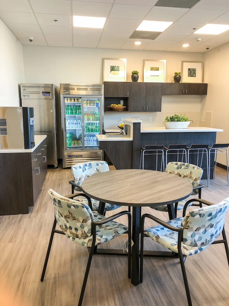 A bright kitchen with a modern countertop, bar seating, and a dining table with patterned chairs. The space includes a fridge, cabinets, and neatly arranged refreshments.