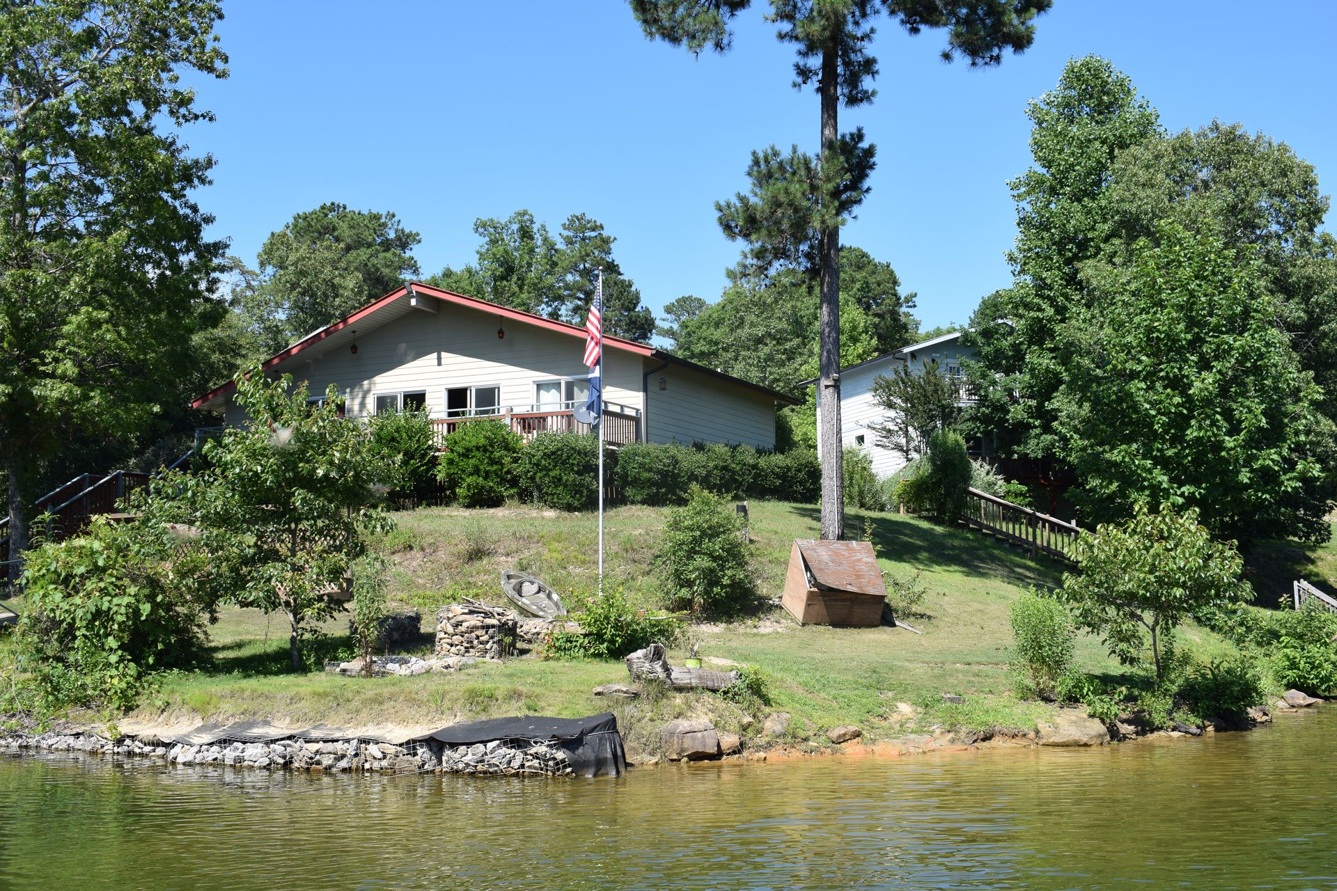 Lakeside residential buildings surrounded by mature trees, with a grassy shoreline, deck, and calm water in the foreground