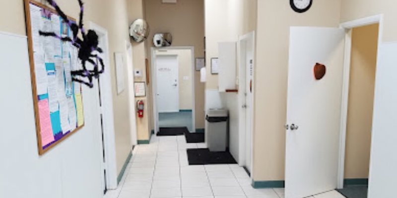  A clean hallway with white tiles and beige walls. A bulletin board on the wall features colorful papers, and several open doors lead to different rooms.