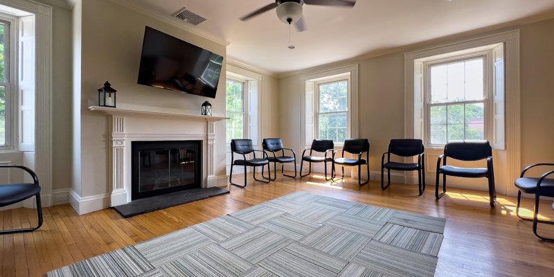 A bright and spacious room with a cozy fireplace, ample seating arranged in a circle, and large windows, creating a warm and inviting space for group activities.