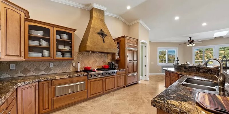  Spacious kitchen with granite countertops, custom wood cabinetry, a large range hood, stainless steel appliances, and ample natural light.