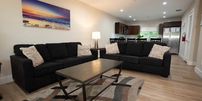 A modern living room with cozy black sofas, a stylish coffee table, and a vibrant wall art, seamlessly connecting to a sleek kitchen area.