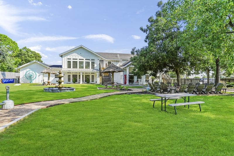 Grassy yard with picnic table, fountain, and shaded seating areas outside a large white house
