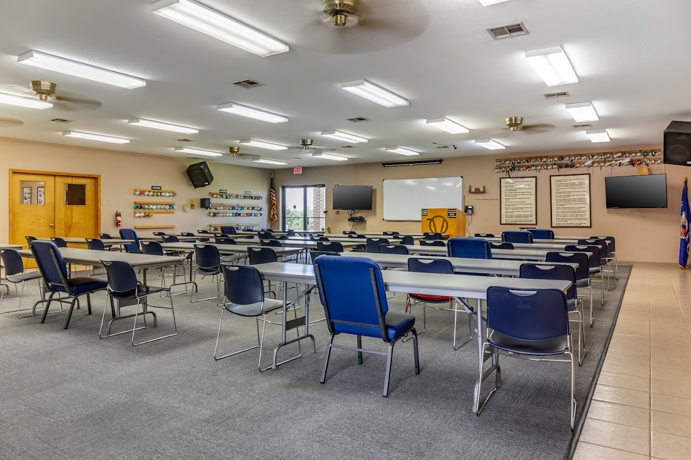 A spacious classroom with rows of tables and chairs, a whiteboard, a podium, and various educational materials on the walls.
