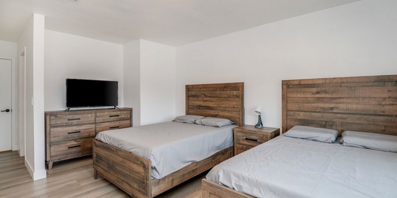 A modern bedroom featuring two rustic wood-framed beds with gray linens, matching nightstands, a wooden dresser, and a sleek flat-screen TV.