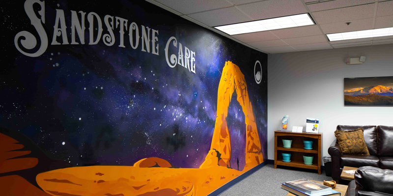 A vibrant mural of a desert arch and starry night sky brings warmth to a cozy waiting area with leather seating and shelves for books.