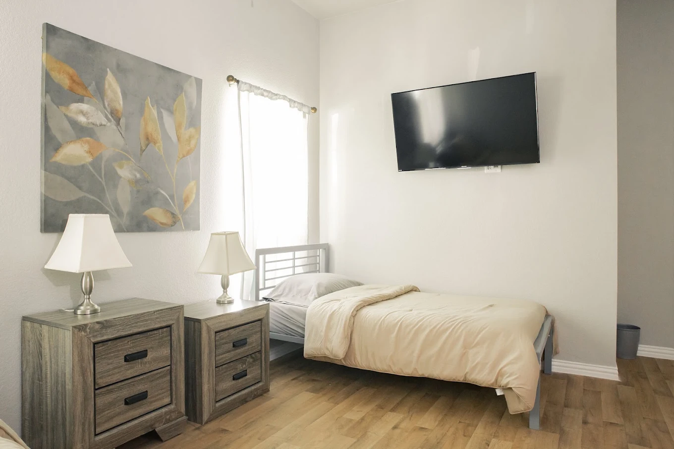 Bright bedroom with a single bed, bedside cabinets, lamps, wall art, wood flooring, and a wall-mounted television