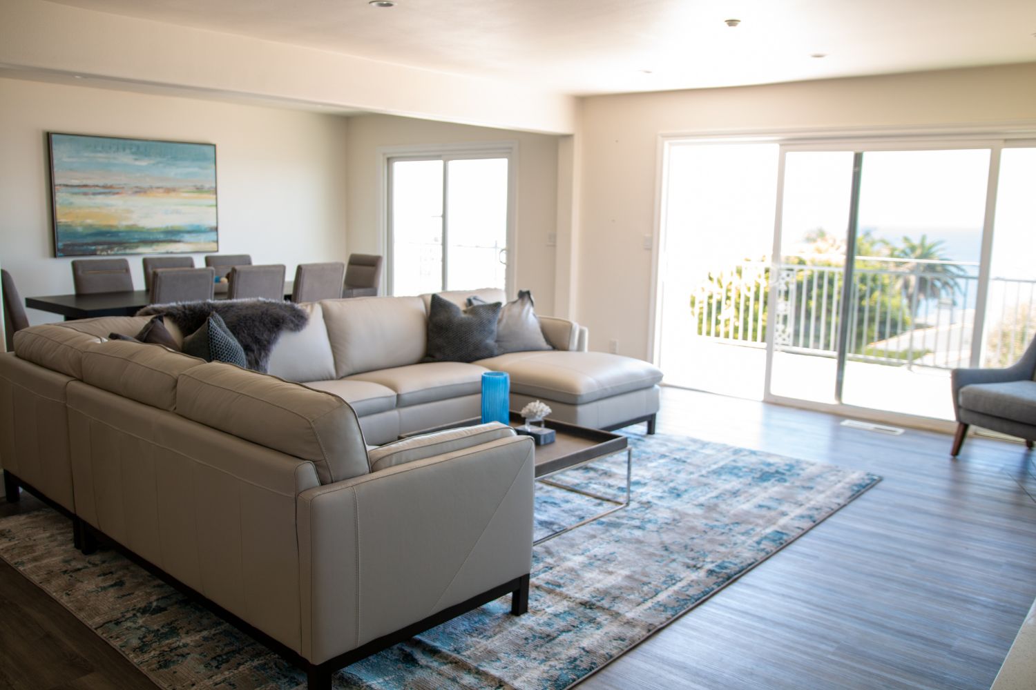 Bright and open living space featuring a modern sectional sofa, large dining table, and expansive ocean view through sliding glass doors.