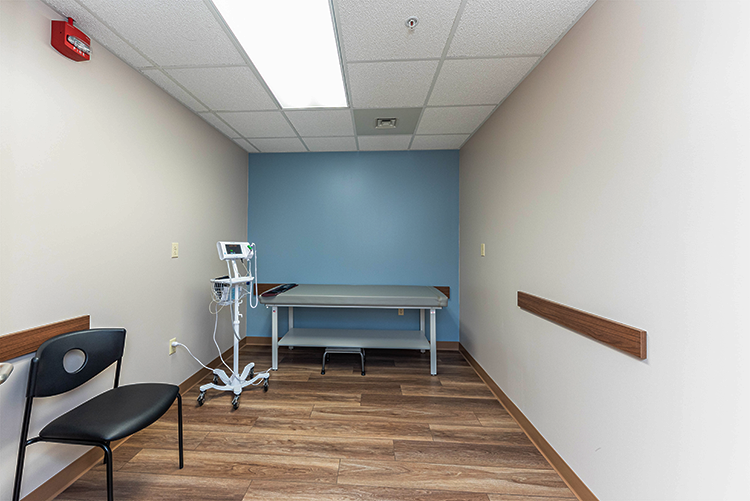 BrightView Health exam room with examination table, vital signs monitor, patient chair, blue accent wall, and wood-look flooring
