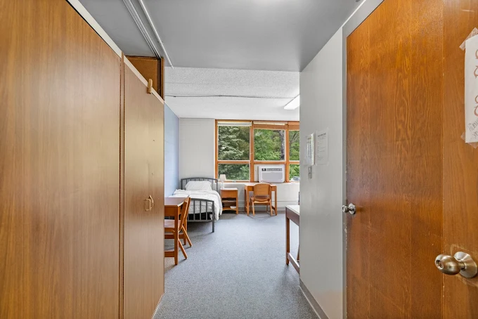 View of a residential bedroom from the doorway with twin beds, desks, and large window overlooking trees.