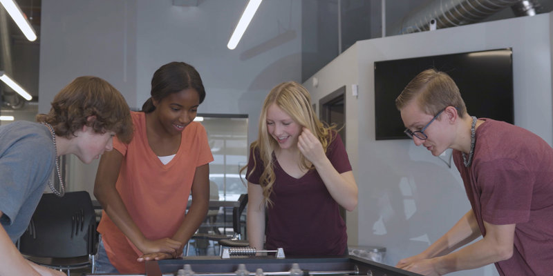 A group of smiling teenagers enjoys a friendly foosball game, creating a fun and engaging environment in a modern, well-lit space.