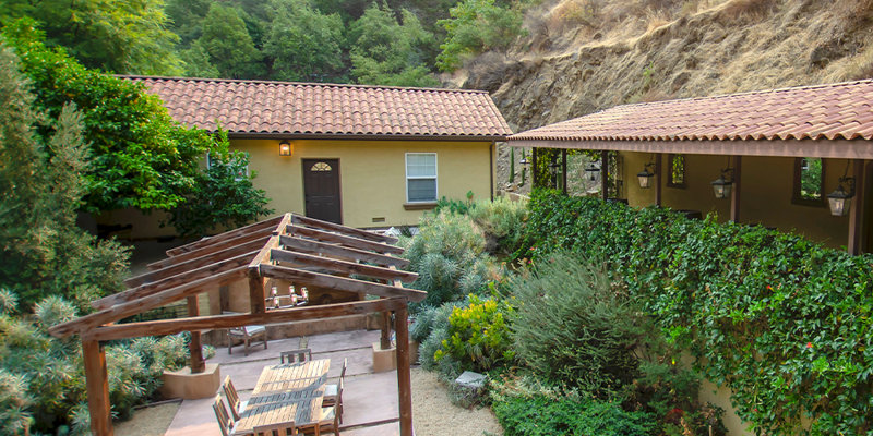 A peaceful outdoor patio with cozy seating, a wooden pergola, and lush greenery, set against a backdrop of scenic hills and natural beauty.