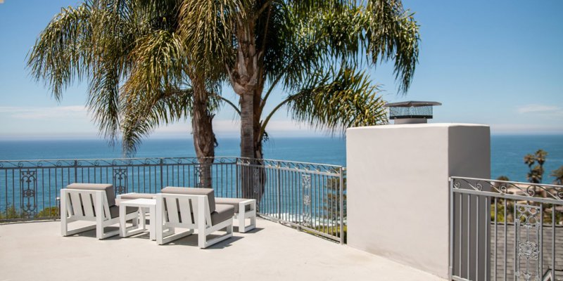 Stunning ocean-view patio with cozy seating, palm trees, and elegant railings, offering a serene and relaxing outdoor space under clear skies.