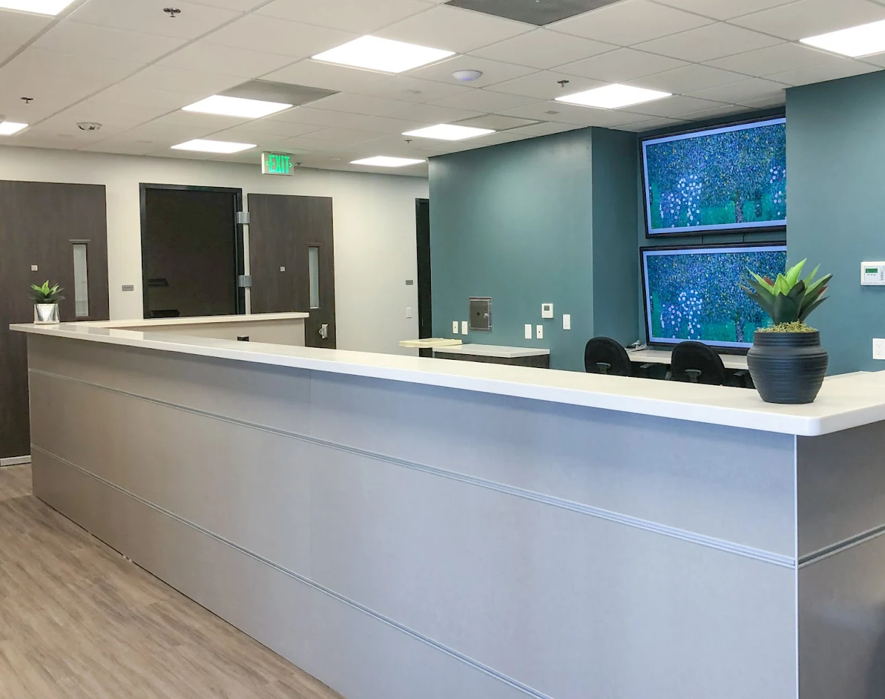 A sleek reception area with a long white counter, potted plants, and dual flat-screen monitors on a blue accent wall behind the desk.