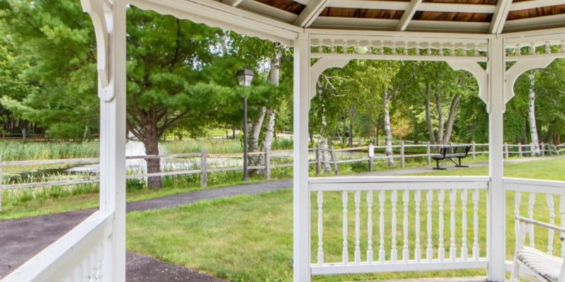 A serene view from a white gazebo overlooking a peaceful path, greenery, and a wooden fence, offering a tranquil outdoor space surrounded by nature.