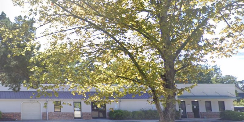 A modest one-story building with large windows, framed by a tree with lush, green leaves, creating a welcoming and tranquil environment.