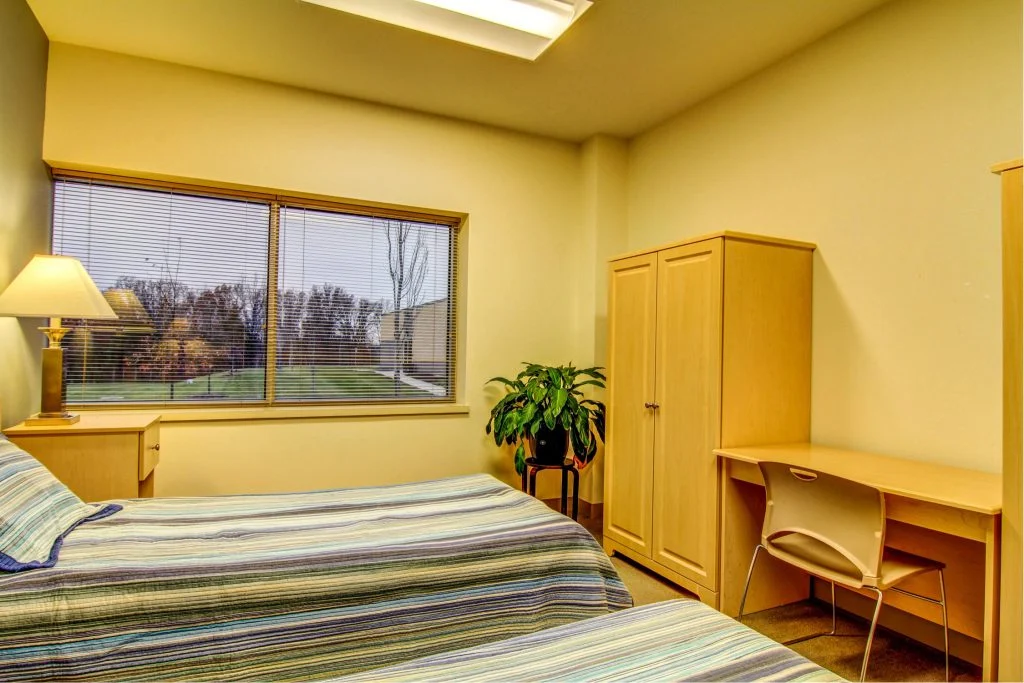Shared patient bedroom with two beds, wardrobe, and desk.