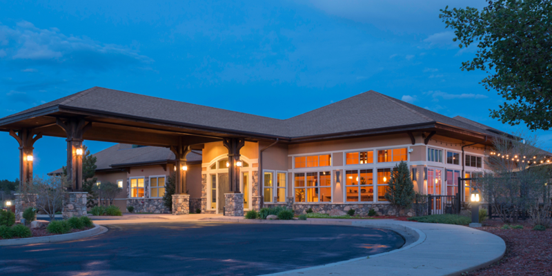 A warmly lit, welcoming building with large windows, surrounded by greenery, creating a serene and inviting atmosphere at dusk.