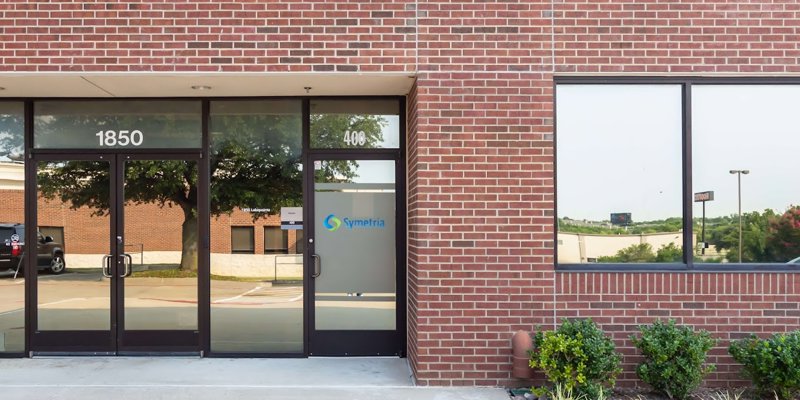  Modern brick office building entrance with large glass doors and windows, surrounded by neatly landscaped greenery, creating a welcoming atmosphere.