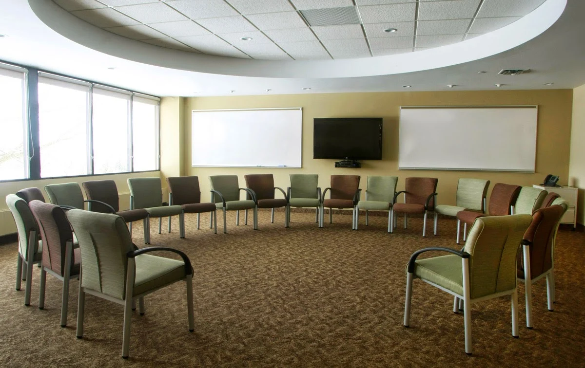 Spacious therapy room with chairs arranged in a circle, large windows, whiteboards, and a wall-mounted television