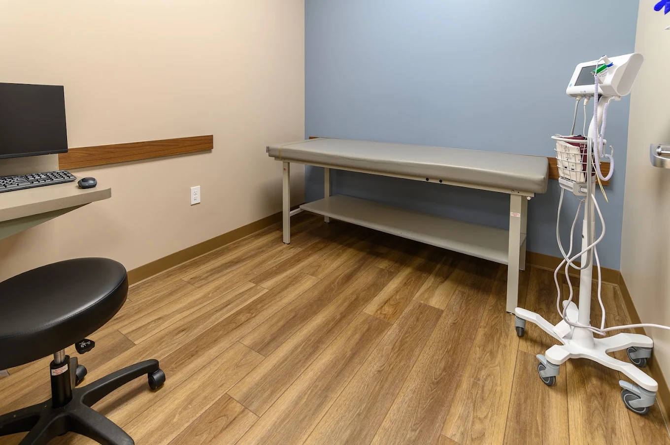 BrightView Health exam room with padded examination table, medical monitoring equipment, computer workstation, rolling stool, blue accent wall, and wood-look flooring