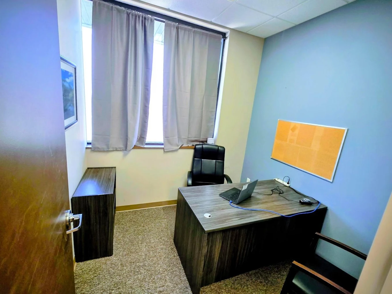 Private consultation room at BrightView Health with desk, laptop, office chair, guest chair, corkboard, curtained window, and blue accent wall