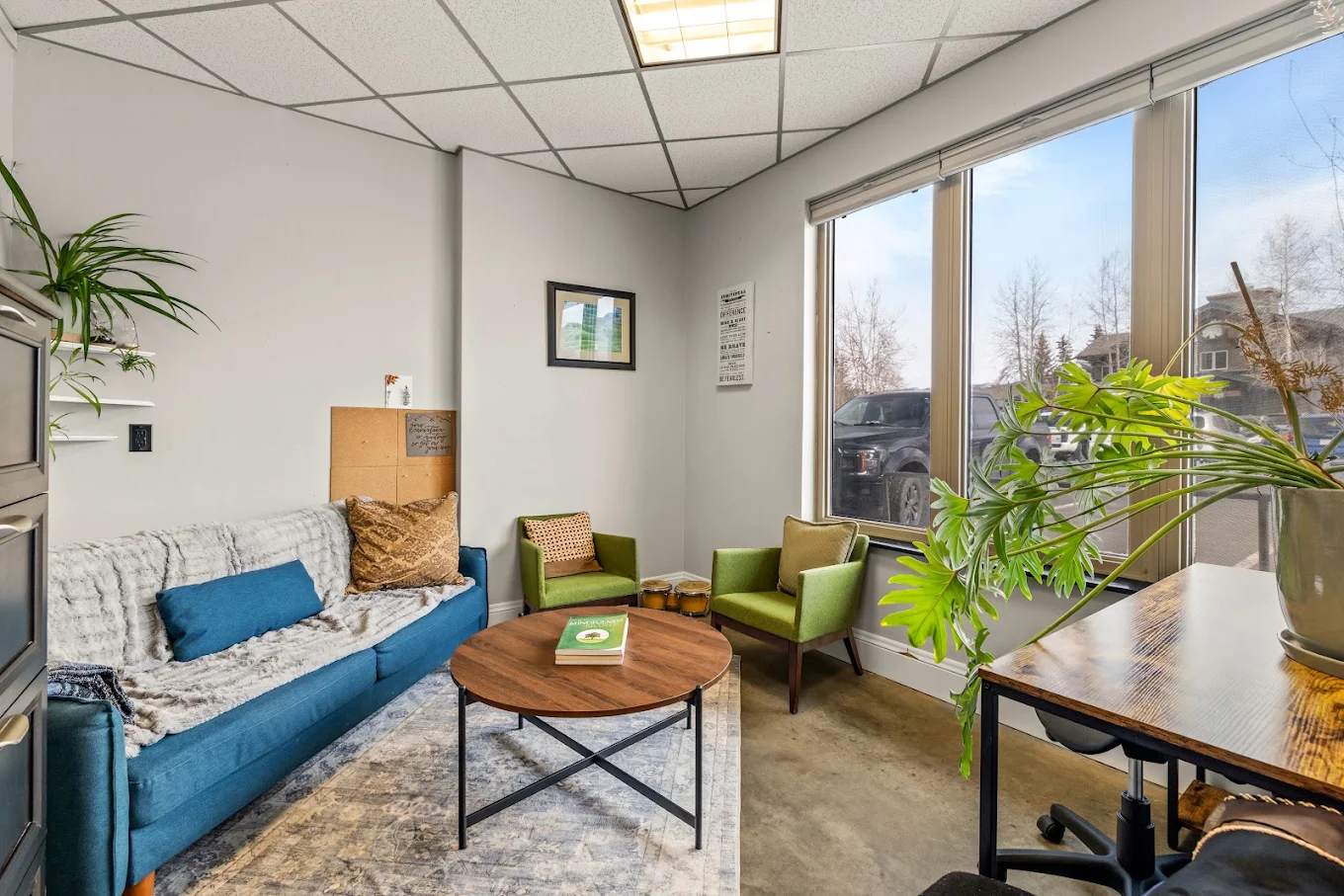 Comfortable counseling room with a blue sofa, green armchairs, coffee table, plants, and large windows