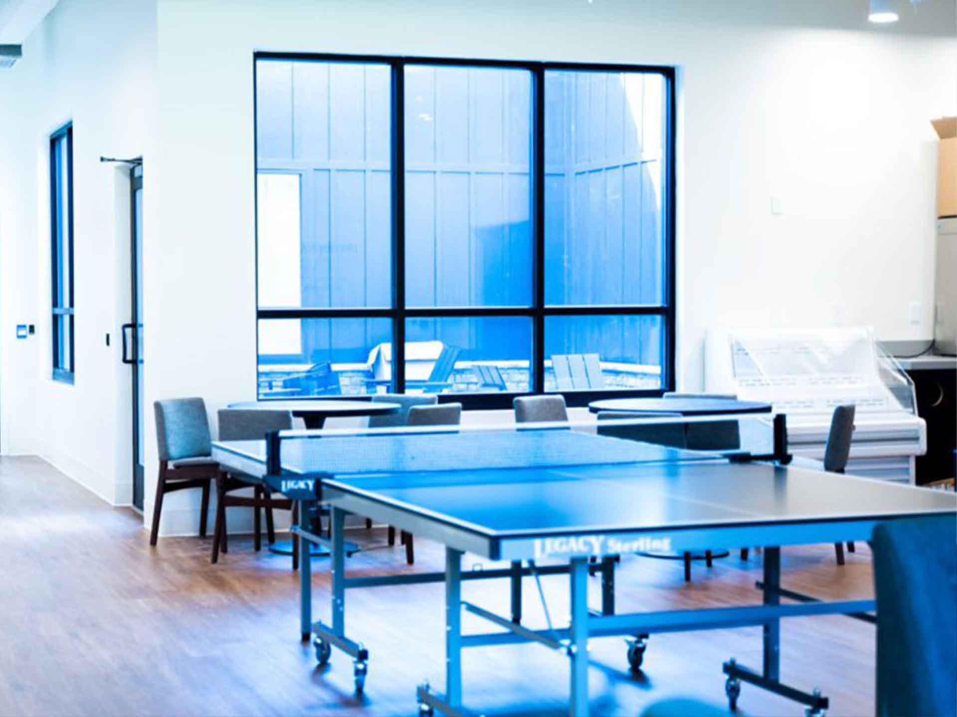 A rec room with a ping-pong table, chairs, and large windows.