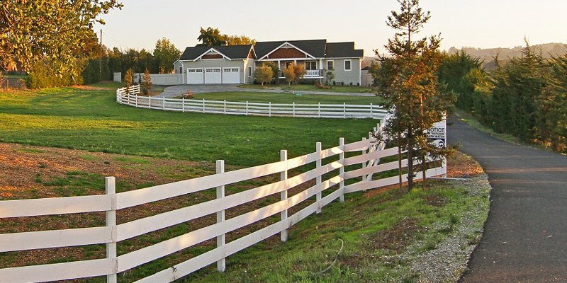 A serene countryside home with a long driveway, surrounded by lush greenery and a charming white picket fence, offering a peaceful retreat.