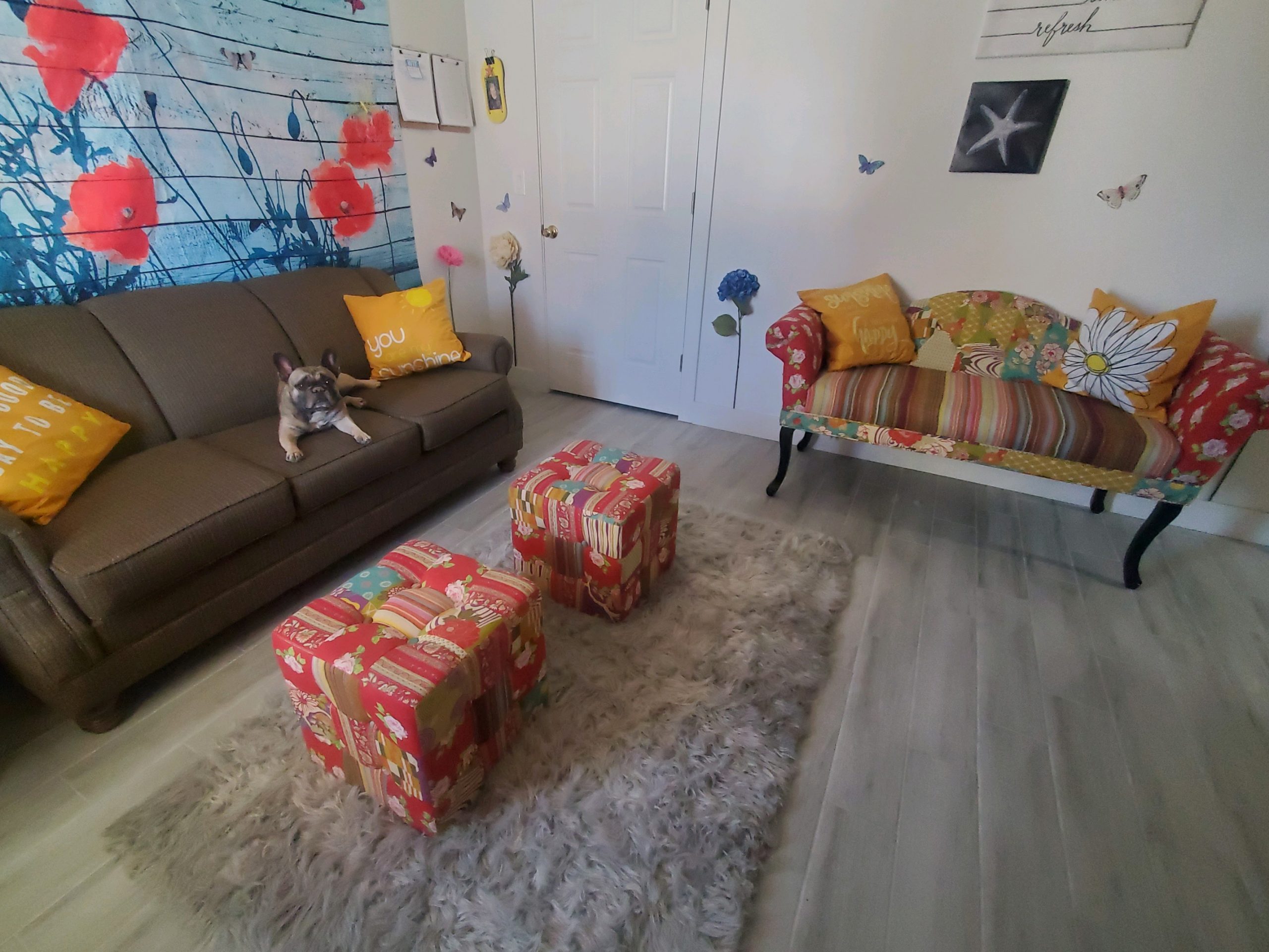 Bright and cheerful sitting room with a tapestry wall, couch, floral loveseat, pillows, and a small dog resting on the sofa