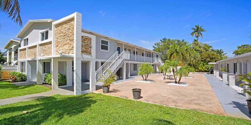 A bright, modern apartment complex features well-kept greenery, palm trees, and a spacious courtyard, creating a welcoming and vibrant atmosphere.