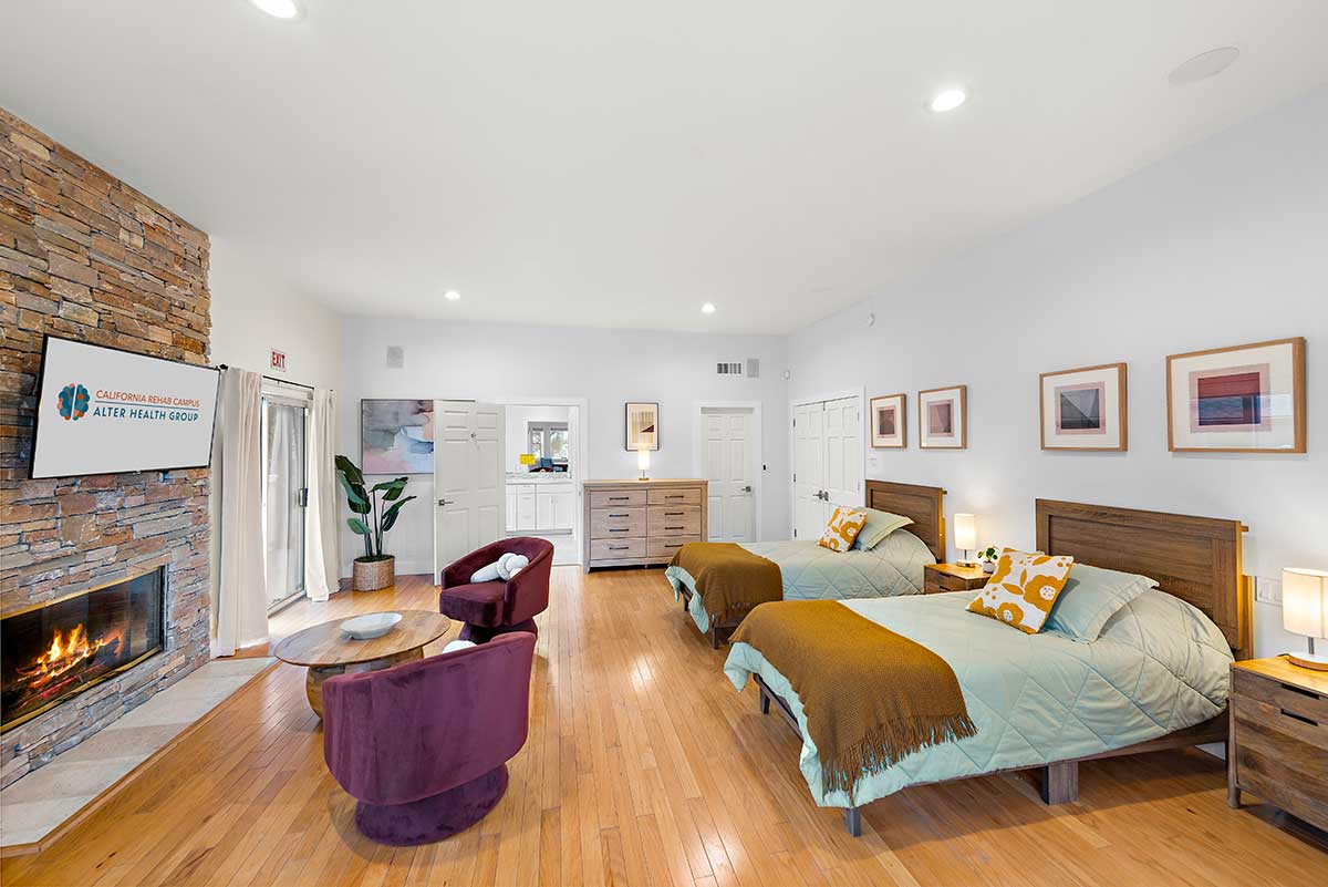 Spacious shared bedroom with two beds, wood flooring, stone fireplace, seating area, dresser, and bedside lamps
