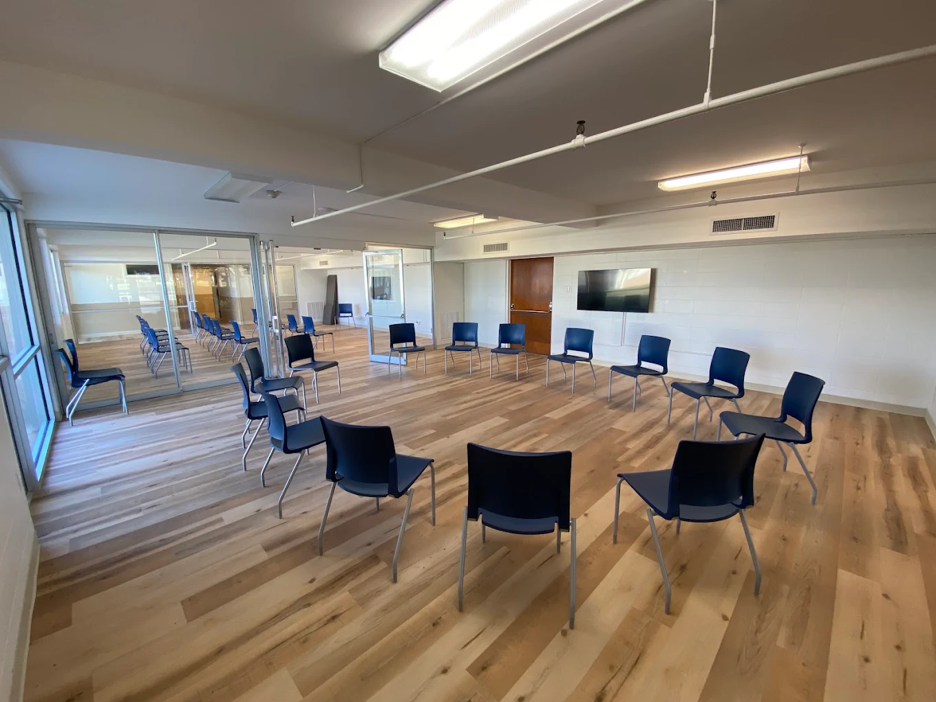 Group therapy room with blue chairs arranged in a circle, wood flooring, glass partitions, and wall-mounted television