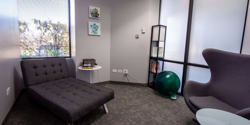 A cozy therapy room with a gray chaise, modern chair, bookshelf, and large windows, creating a peaceful and relaxing atmosphere