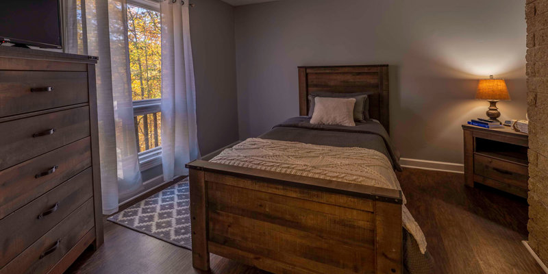 Cozy bedroom with a rustic wooden bed and dresser, warm lighting, and a window offering a peaceful view of autumn trees outside.