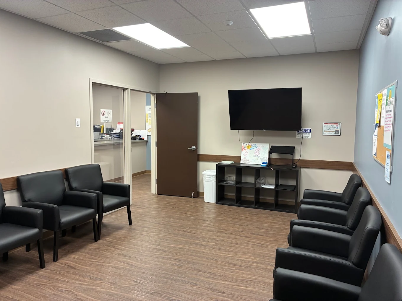 Treatment room with black armchairs arranged along the walls, wall-mounted television, shelving, and bulletin board
