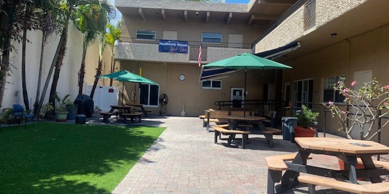 A sunny outdoor courtyard with picnic tables, green umbrellas, and lush palm trees, providing a comfortable and relaxing space for gatherings.