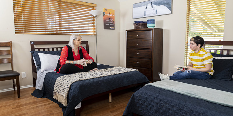 Two women sit on twin beds in a bright, cozy bedroom with warm wooden floors and soft lighting, engaged in a thoughtful conversation.