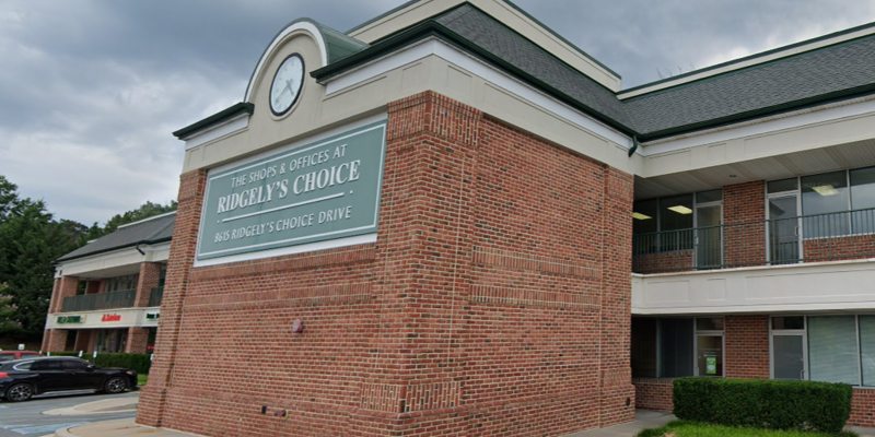  A two-story brick building with a prominent sign reading "Ridgely's Choice." A large clock is mounted above the entrance, with storefronts below.