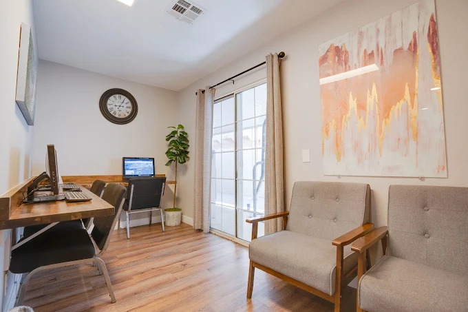 Small computer room with wooden desks, computers, wall clock, and gray chairs for study or work.