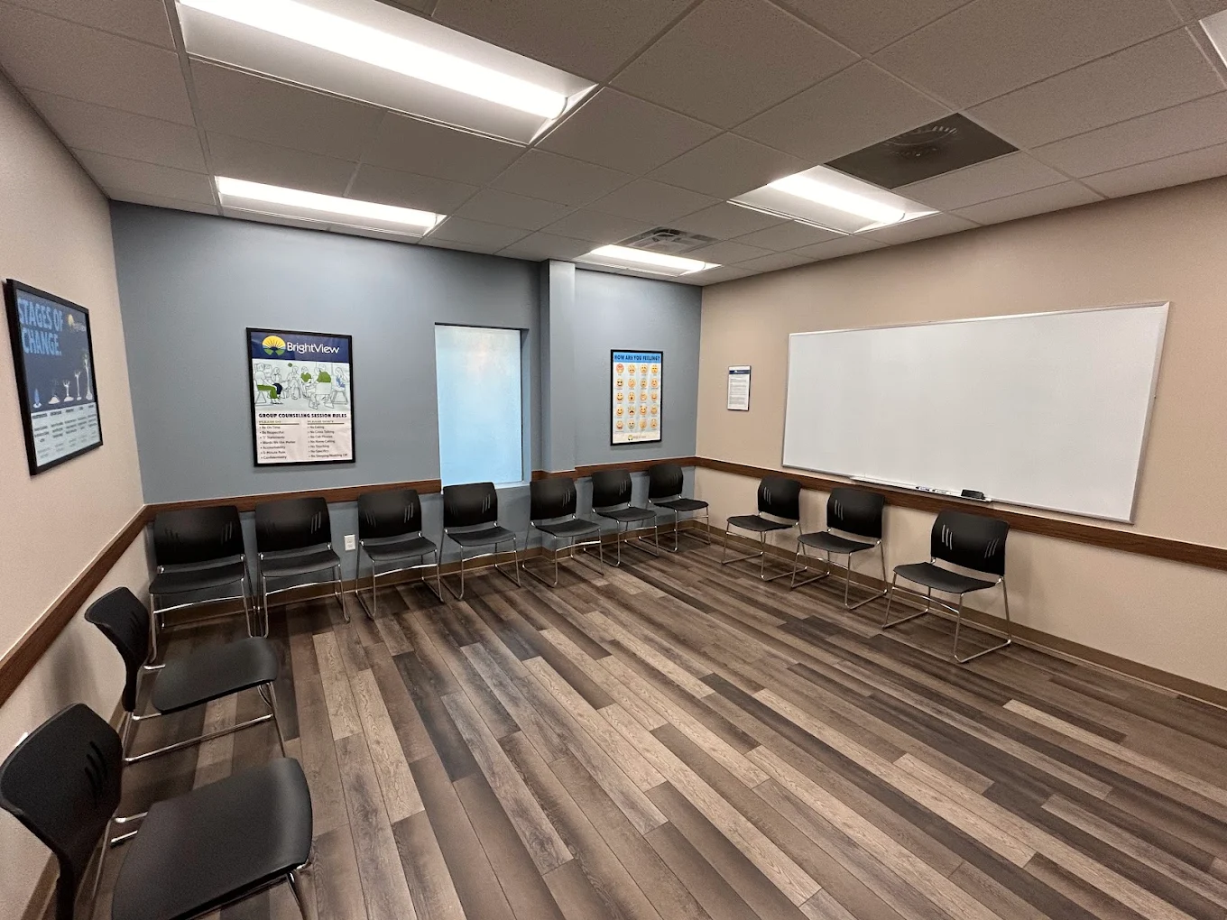 BrightView Health group counseling room with black chairs arranged around the walls, large whiteboard, recovery posters, blue accent wall, and wood-look flooring