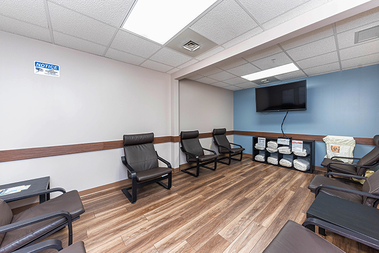 BrightView Health treatment room with cushioned chairs arranged around the walls, wall-mounted TV, storage shelves, blue accent wall, and wood-look flooring