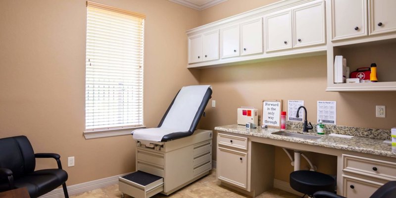  Welcoming exam room with a comfortable patient bed, warm beige walls, ample cabinetry, and natural light from a large window.