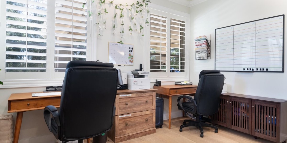 Office space with two wooden desks, black chairs, and large windows with plantation shutters