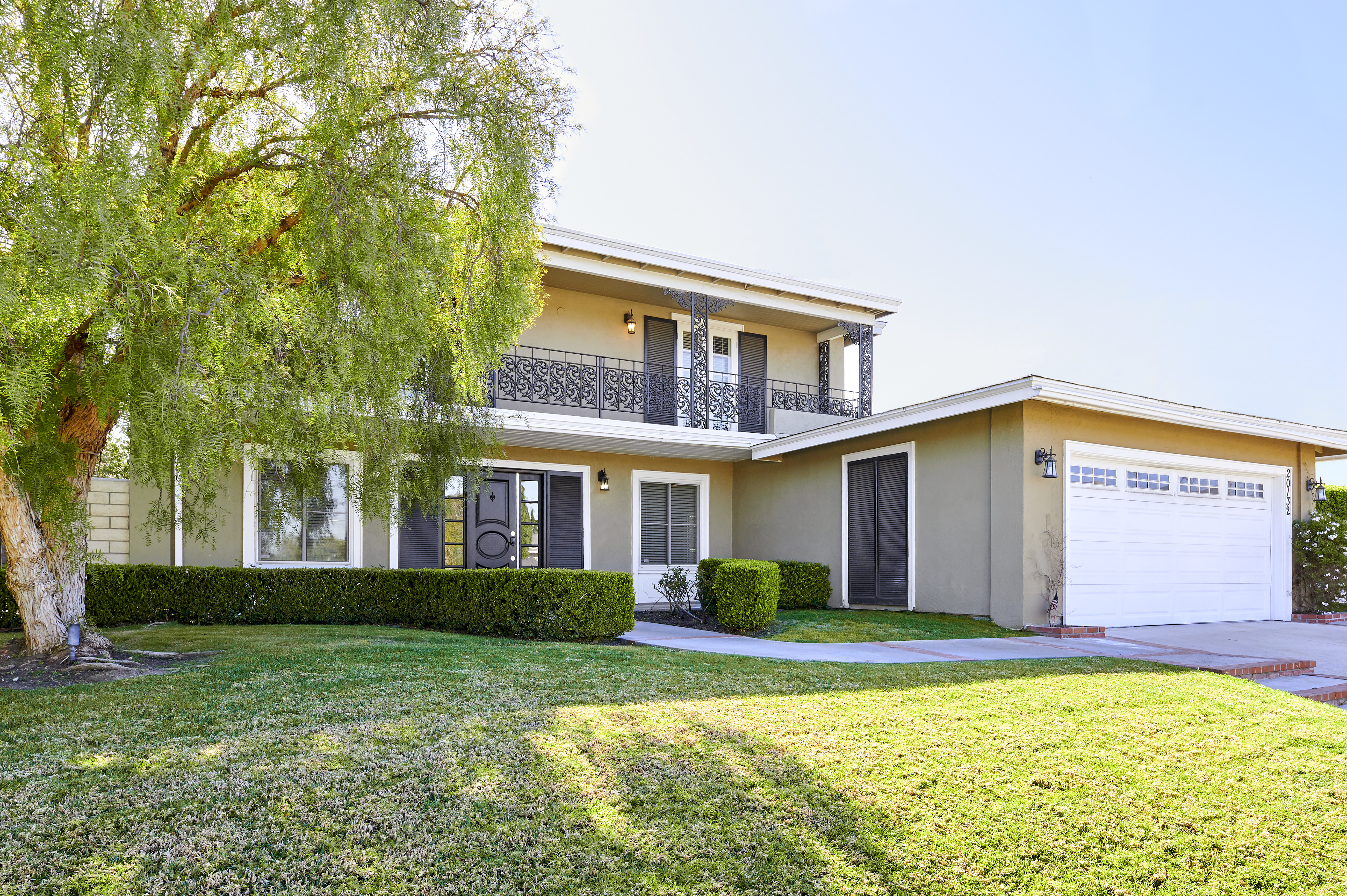 A charming two-story home with a balcony, decorative iron railings, and a well-kept lawn, shaded by a large tree in a peaceful setting.
