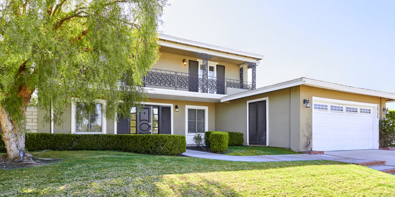 A charming two-story home with a balcony, decorative iron railings, and a well-kept lawn, shaded by a large tree in a peaceful setting.