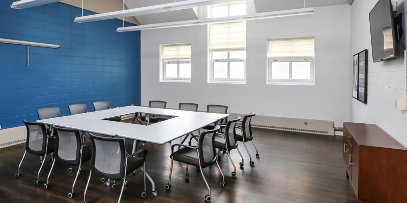  Bright, modern meeting room with large windows, blue accent wall, and a spacious conference table surrounded by comfortable chairs on wheels.